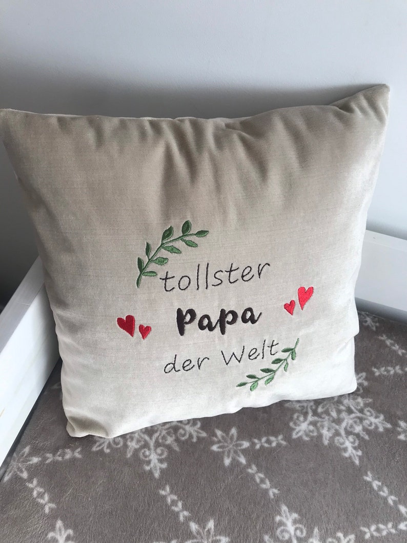 May include: Beige throw pillow with embroidered green leaves and red hearts. The text "tollster Papa der Welt" is embroidered in brown on the pillow.