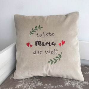 May include: Beige throw pillow with embroidered green leaves and red hearts. The text "tolle Mama der Welt" is embroidered in black.
