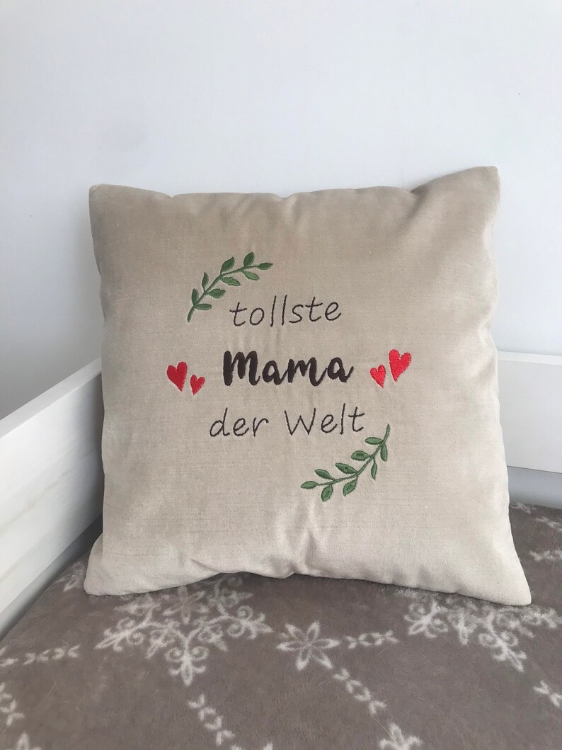 May include: Beige throw pillow with embroidered green leaves and red hearts. The text "tolle Mama der Welt" is embroidered in black.