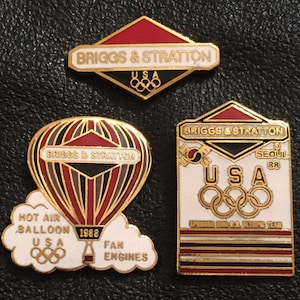 May include: Three gold-colored pins with red, white, and blue accents. The top pin features the text "BRIGGS & STRATTON" and the Olympic rings. The middle pin features a hot air balloon with the text "BRIGGS & STRATTON" and "HOT AIR BALLOON 1988 USA". The bottom pin features the text "BRIGGS & STRATTON" and "SEOUL 88 USA" with the Olympic rings and the text "SPIN88 U.S.A. OLYMPIC TEAM".
