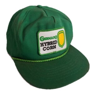 May include: Green baseball cap with a white patch that says "Germain's Hybrid Corn" in black and green letters. The patch has a yellow corn kernel graphic.