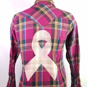 May include: A red, black, blue, and brown plaid flannel shirt with a white ribbon design on the back.