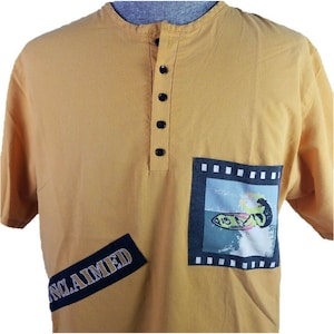 Pode incluir: Uma camisa amarela de manga curta com gola de botões e um design de tira de filme preto e branco na frente. O design da tira de filme apresenta uma imagem de desenho animado de uma pessoa surfando. A camisa também tem um remendo preto com a palavra "UNCLAIMED" nele.
