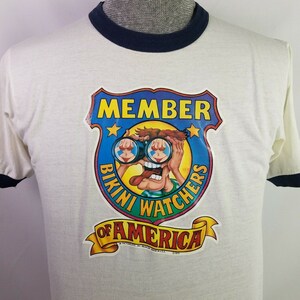 Pode incluir: Camiseta branca com gola azul. A camiseta apresenta um gráfico vintage de um personagem de desenho animado usando binóculos e olhando para um biquíni. O gráfico é um escudo azul e amarelo com o texto "MEMBER BIKINI WATCHERS OF AMERICA".