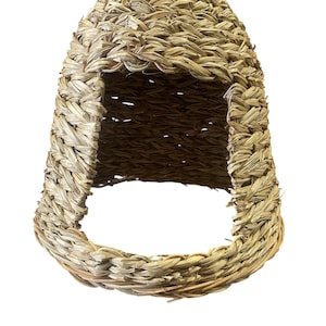 May include: A woven pet house, dome-shaped, crafted from natural, light brown fibres. It has an open front and a rounded entrance, offering a cosy, enclosed space for small animals. The texture of the material is clearly visible.