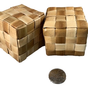May include: Two woven cube-shaped boxes made from light brown and tan natural fibers. The boxes have a checkerboard pattern. A coin is in the foreground for size comparison. The boxes are likely for storage or decorative purposes.