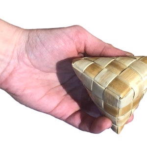 May include: A woven triangular basket made of light brown and beige natural fibers. The basket has a checkered pattern and is held in a hand. The basket is likely handmade and could be used for storage or decoration.
