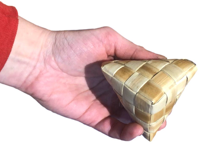 May include: A woven triangular basket made of light brown and beige natural fibers. The basket has a checkered pattern and is held in a hand. The basket is likely handmade and could be used for storage or decoration.