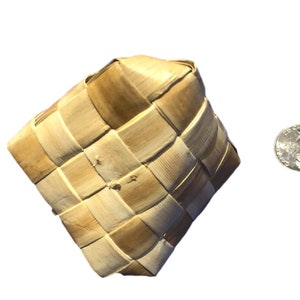 May include: A woven square basket made of light brown natural fibers. The basket has a diamond shape and is next to a US quarter coin for size comparison. The basket is likely handmade and has a rustic appearance.
