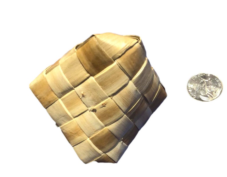 May include: A woven square basket made of light brown natural fibers. The basket has a diamond shape and is next to a US quarter coin for size comparison. The basket is likely handmade and has a rustic appearance.