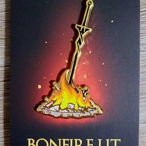 Op de afbeelding: Een gouden emaille pin met daarop een zwaard dat in een brandend kampvuur is gestoken. De pin heeft een zwarte achtergrond en de tekst "BONFIRE LIT" in gouden letters.