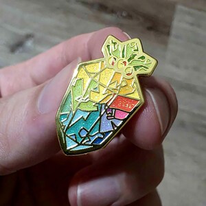 K&ouml;nnte beinhalten: Ein farbenfroher Emaille-Pin in Form eines Geschenkbeutels. Der Pin hat einen goldenen Umriss und ist mit einem Mosaik aus Regenbogenfarben gef&uuml;llt, darunter Gr&uuml;n, Gelb, Blau und Rosa. Die Oberseite des Pins hat ein sternf&ouml;rmiges Design.