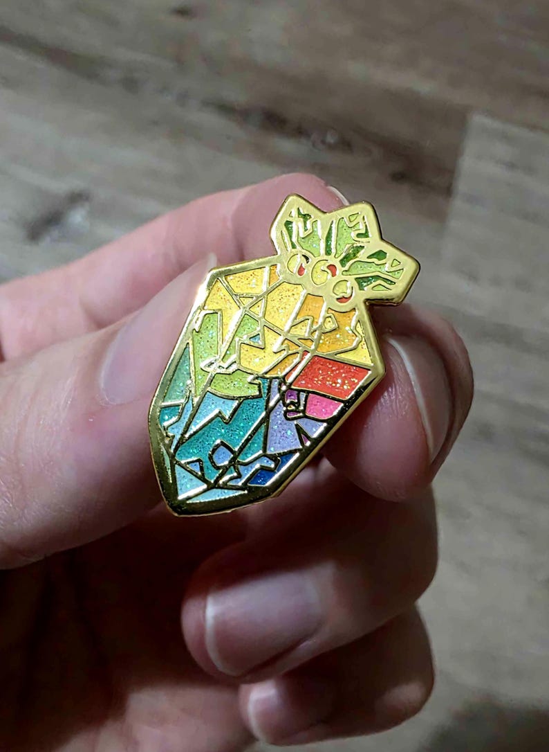 K&ouml;nnte beinhalten: Ein farbenfroher Emaille-Pin in Form eines Geschenkbeutels. Der Pin hat einen goldenen Umriss und ist mit einem Mosaik aus Regenbogenfarben gef&uuml;llt, darunter Gr&uuml;n, Gelb, Blau und Rosa. Die Oberseite des Pins hat ein sternf&ouml;rmiges Design.