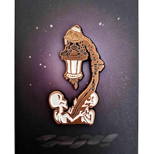 May include: A brown and white enamel pin depicting a stylized lantern with two skeleton figures. The lantern has a white light and detailed design. The pin is set against a dark background with a starry pattern.