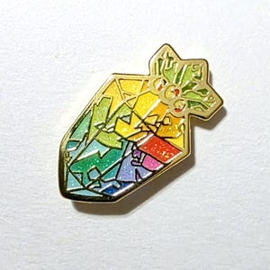 K&ouml;nnte beinhalten: Ein goldfarbener Emaille-Pin in Form eines stilisierten Edelsteins. Der Edelstein ist in Abschnitte in Regenbogenfarben unterteilt, darunter Rot, Orange, Gelb, Gr&uuml;n, Blau und Lila. Die Oberseite des Pins hat ein sternf&ouml;rmiges Design mit gr&uuml;nen Akzenten.
