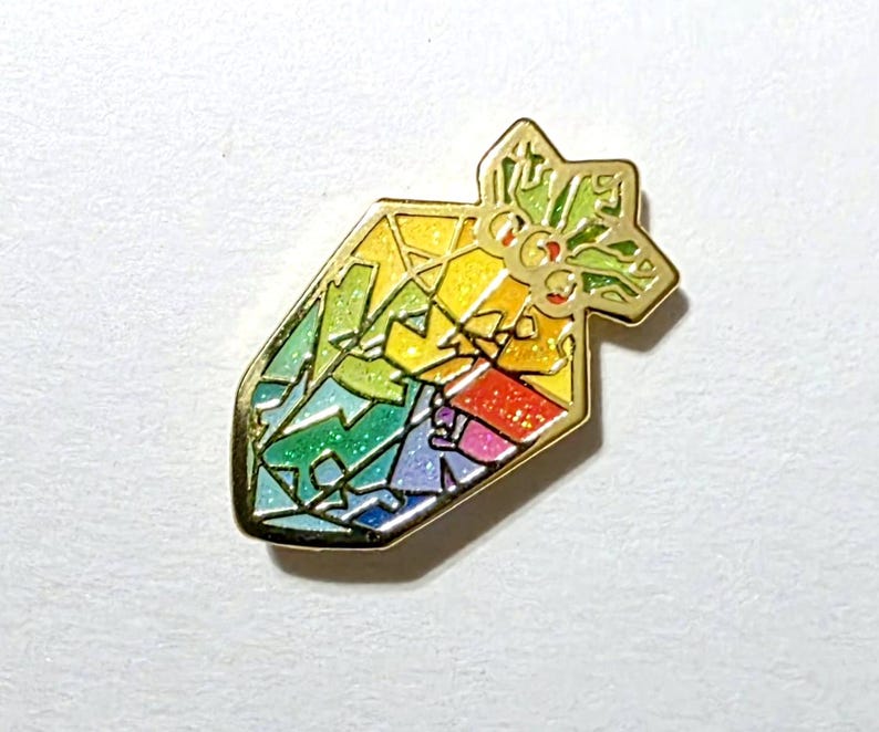 K&ouml;nnte beinhalten: Ein goldfarbener Emaille-Pin in Form eines stilisierten Edelsteins. Der Edelstein ist in Abschnitte in Regenbogenfarben unterteilt, darunter Rot, Orange, Gelb, Gr&uuml;n, Blau und Lila. Die Oberseite des Pins hat ein sternf&ouml;rmiges Design mit gr&uuml;nen Akzenten.