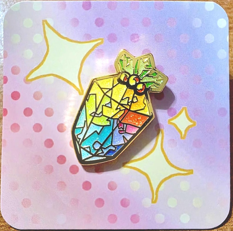 K&ouml;nnte beinhalten: Ein farbenfroher Emaille-Pin mit einem Buntglas-Design. Der Pin hat eine goldene Umrandung und ein geometrisches Muster aus Gelb, Blau, Gr&uuml;n und Rot. Der Pin befindet sich auf einem rosa-wei&szlig; gepunkteten Hintergrund mit Sternen-Akzenten.