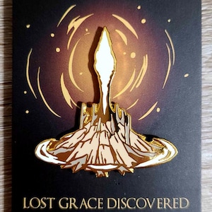 Op de afbeelding: Gouden emaille pin met een gestileerde afbeelding van een gouden toren die uit een rotsachtige basis oprijst. De toren wordt omringd door een wervelende, gouden halo. De tekst "LOST GRACE DISCOVERED" is onder de afbeelding gedrukt.