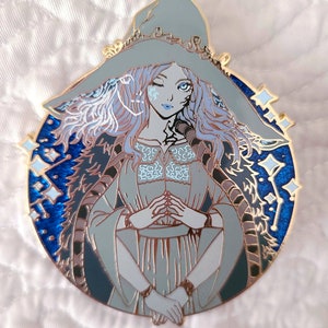 May include: A detailed enamel pin featuring a fantasy character with blue skin, wearing a large hat and a flowing gown. The pin has a gold border and a blue background with star-like accents. The character has long, wavy, light blue hair.