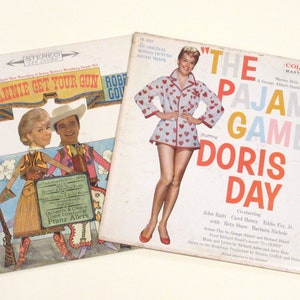 May include: Two vintage vinyl record albums featuring Doris Day. The first album is "Annie Get Your Gun" with a colourful illustration of Doris Day in a cowboy outfit. The second album is "The Pajama Game" with a photo of Doris Day in a white and red heart-patterned pyjama top and shorts.