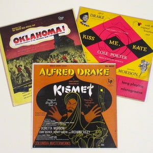 May include: Three vintage vinyl record covers. The top cover is yellow with red and black accents and features the text "Kiss Me, Kate" and "Cole Porter". The middle cover is orange and black and features the text "Alfred Drake" and "Kismet". The bottom cover is red, yellow, and black and features the text "Oklahoma!" and "The Theatre Guild Musical Play".