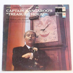 May include: A vintage vinyl record cover for Captain Kangaroo's Treasure House featuring Bob Keeshan in a dark suit standing before a large grandfather clock with a stylized face on the clock dial. The text includes the show title and credits.