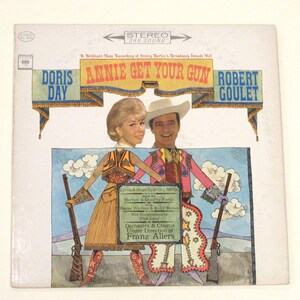 May include: A vintage vinyl record cover featuring Doris Day and Robert Goulet in a colourful illustration for the musical "Annie Get Your Gun". The cover features a bright blue sky with white clouds, and the title of the musical is in large, bold letters.