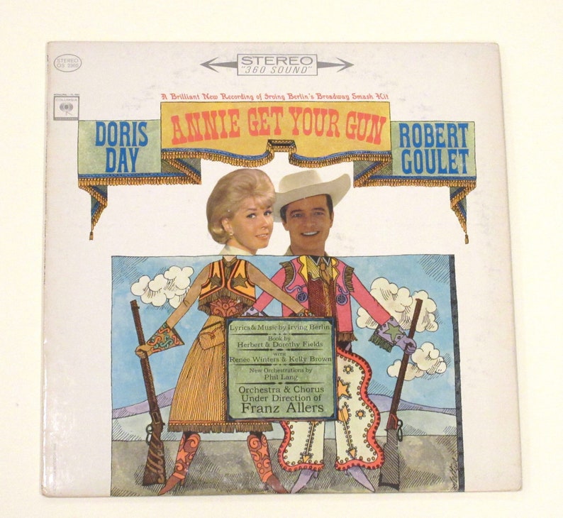 May include: A vintage vinyl record cover featuring Doris Day and Robert Goulet in a colourful illustration for the musical "Annie Get Your Gun". The cover features a bright blue sky with white clouds, and the title of the musical is in large, bold letters.