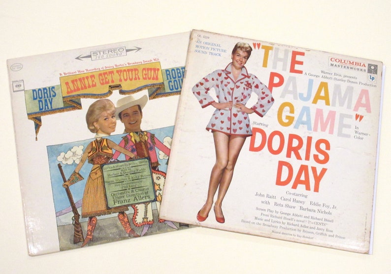 May include: Two vintage vinyl record albums featuring Doris Day. The first album is "Annie Get Your Gun" with a colourful illustration of Doris Day in a cowboy outfit. The second album is "The Pajama Game" with a photo of Doris Day in a white and red heart-patterned pyjama top and shorts.