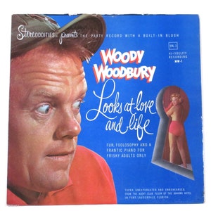May include: A vintage record album cover featuring a man in a red shirt and a baseball cap looking to the right. The album cover is blue and features the text "Woody Woodbury Looks at Love and Life" in a white cursive font. The album cover also features the text "StereoDITIES Presents The Party Record With A Built-In Blush" in a white cursive font. The album cover also features the text "Vol. 1 Hi-Fidelity Recording MW-1" in a white sans-serif font.