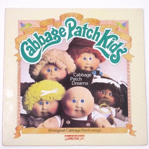 May include: A vintage Cabbage Patch Kids record album cover featuring five Cabbage Patch Kids dolls with the title "Cabbage Patch Kids" and the subtitle "Cabbage Patch Dreams". The album cover is pink and white with green text and a yellow ribbon.
