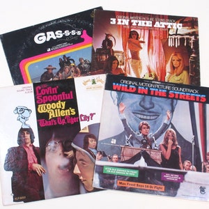 May include: Four vinyl record albums with different covers. The top album is black with yellow, orange, and red stripes and the text "GAS-S-SS". The album below it is red, white, and blue with the text "3 IN THE ATTIC". The album on the left is white with a cartoon character and the text "Lovin Spoonful Woody Allen's What's Up, Tiger Lily?". The album on the right is blue with the text "WILD IN THE STREETS".