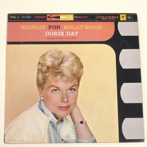 May include: A vintage vinyl record album cover featuring a portrait of Doris Day, a blonde woman wearing a white sweater and a blue shirt. The album title is "Hooray for Hollywood" and the record label is Columbia.