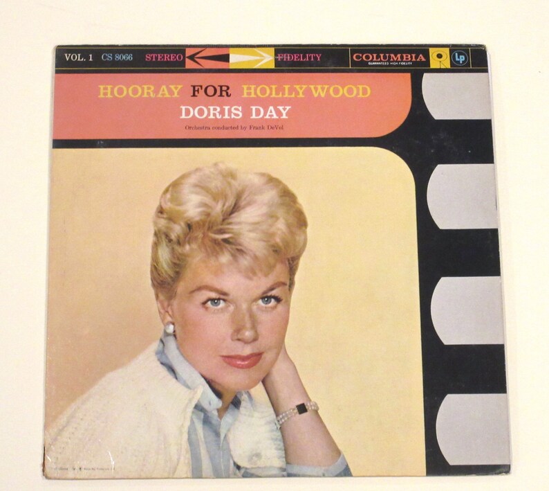 May include: A vintage vinyl record album cover featuring a portrait of Doris Day, a blonde woman wearing a white sweater and a blue shirt. The album title is "Hooray for Hollywood" and the record label is Columbia.