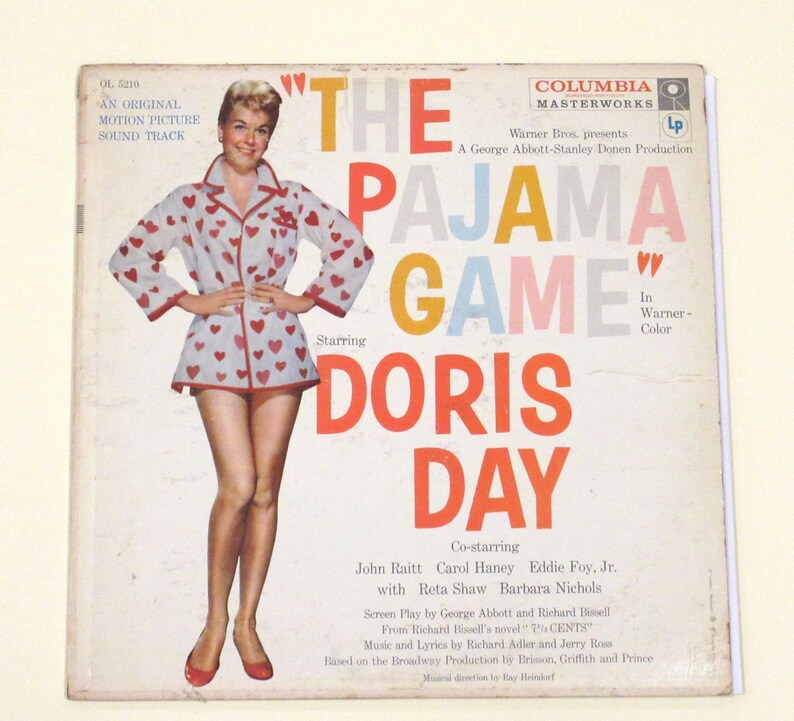 May include: A vintage Columbia Masterworks LP record cover featuring Doris Day in a white and red heart-patterned pyjama top and red shoes. The title of the record is "The Pajama Game" in large, colourful letters. The cover also includes the names of the co-stars, John Raitt, Carol Haney, Eddie Foy Jr., Reta Shaw, and Barbara Nichols.