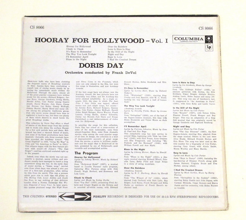 May include: A vintage vinyl record cover for "Hooray for Hollywood - Vol. I" by Doris Day. The cover features a photo of Doris Day in a black and white dress, holding a microphone. The album cover is in a light blue and white colour scheme, with the title of the album in large, bold letters. The album cover also includes a list of songs on the album, including "It's Easy to Remember", "The Way You Look Tonight", and "Over the Rainbow".