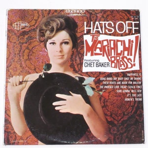 May include: A vintage vinyl record cover featuring a woman holding a black hat. The cover is red and gold with the text "Hats Off The Mariachi Brass! Featuring Chet Baker" and a list of song titles.