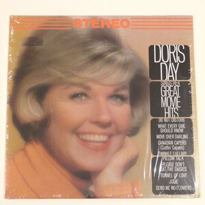 May include: A vintage vinyl record album cover featuring Doris Day, a famous American actress and singer. The cover is orange and white with a photo of Doris Day smiling. The album title is "Doris Day Sings Her Great Movie Hits" and lists the songs included on the record: "Do Not Disturb", "What Every Girl Should Know", "Move Over Darling", "Canadian Capers", "Twinkle Lullaby", "Pillow Talk", "Please Don't Eat The Daisies", "Tunnel Of Love", "Julie", and "Send Me No Flowers."