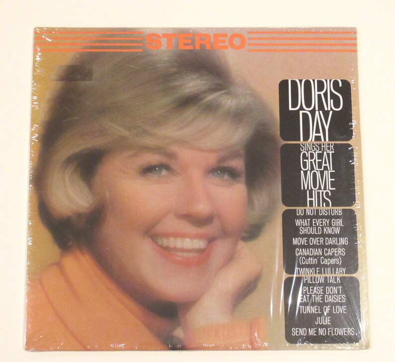 May include: A vintage vinyl record album cover featuring Doris Day, a famous American actress and singer. The cover is orange and white with a photo of Doris Day smiling. The album title is "Doris Day Sings Her Great Movie Hits" and lists the songs included on the record: "Do Not Disturb", "What Every Girl Should Know", "Move Over Darling", "Canadian Capers", "Twinkle Lullaby", "Pillow Talk", "Please Don't Eat The Daisies", "Tunnel Of Love", "Julie", and "Send Me No Flowers."