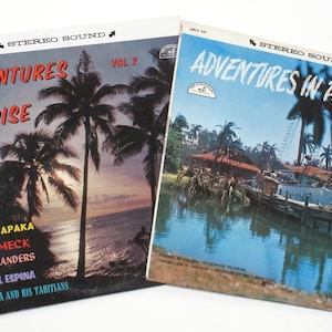 May include: Two vintage vinyl records with a tropical theme. The covers feature palm trees, a sunset, and a boat in a lagoon. The text on the covers reads "Adventures in Paradise" and lists the artists: Alfred Apaka, Roy Smeck, The Islanders, Angel Espina, and Terorotua and His Tahitians.