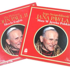 May include: Two red vinyl record albums featuring portraits of Pope John Paul II. The album on the left is titled "His Holiness John Paul II Mass In English". The album on the right is titled "Jego Swiątobliwość Jana Pawła II Msza W Języku Polskim".