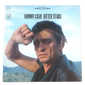May include: Album cover for Johnny Cash's "Bitter Tears." The album art features a portrait of Johnny Cash with a serious expression. The album title and artist name are printed in bold, black letters against a light blue background.