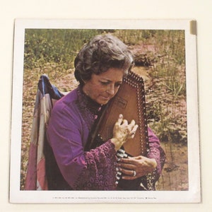 May include: A woman in a purple dress with lace trim plays a dulcimer. She is sitting outdoors in a grassy area.