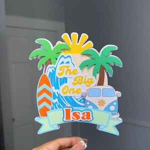 Pode incluir: Um topo de bolo de papel multicamadas com um sol amarelo, palmeiras verdes, uma onda azul, uma prancha de surf laranja e uma carrinha branca e azul. O texto diz The Big One e o nome Isa está num banner verde claro na parte inferior.