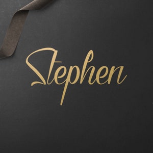 May include: Gold script lettering of the name "Stephen" on a dark grey background. A dark brown ribbon and a small black box with gold-coloured accessories are also present. The overall aesthetic is elegant and minimalist.