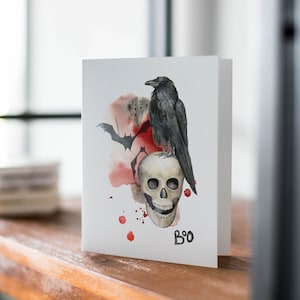 May include: A white card with a watercolor illustration of a black raven perched on a white skull. The raven has a black beak and black eyes. The skull has a toothy grin. There are red watercolor splatters around the raven and skull. The card says "Boo" in black lettering at the bottom.