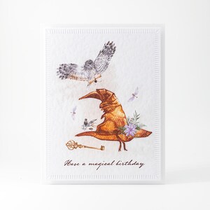 May include: A white card with a watercolor illustration of a brown wizard's hat, a white owl, and a golden key. The text "Have a magical birthday" is printed on the card.