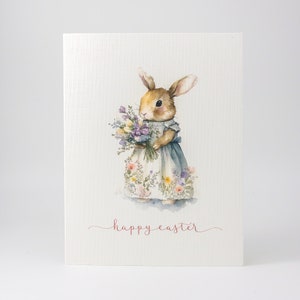 May include: A watercolor illustration of a brown bunny rabbit wearing a blue and white floral dress and holding a bouquet of purple and white flowers. The text "happy easter" is written in pink below the bunny.