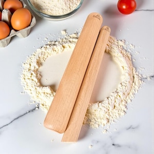 Puede incluir: Dos rodillos de madera con la marca "EUcook" se encuentran sobre una superficie de mármol blanco espolvoreada con harina. También se ven una caja de huevos, un cuenco de harina y un tomate, lo que sugiere un tema de repostería.