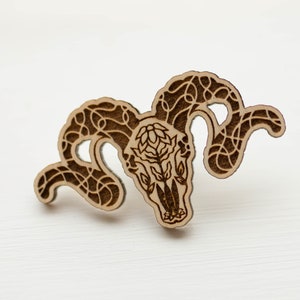 May include: A wooden cutout of a ram's skull with intricate floral designs carved into the horns and skull. The cutout is a light brown color and has a rustic, natural look.